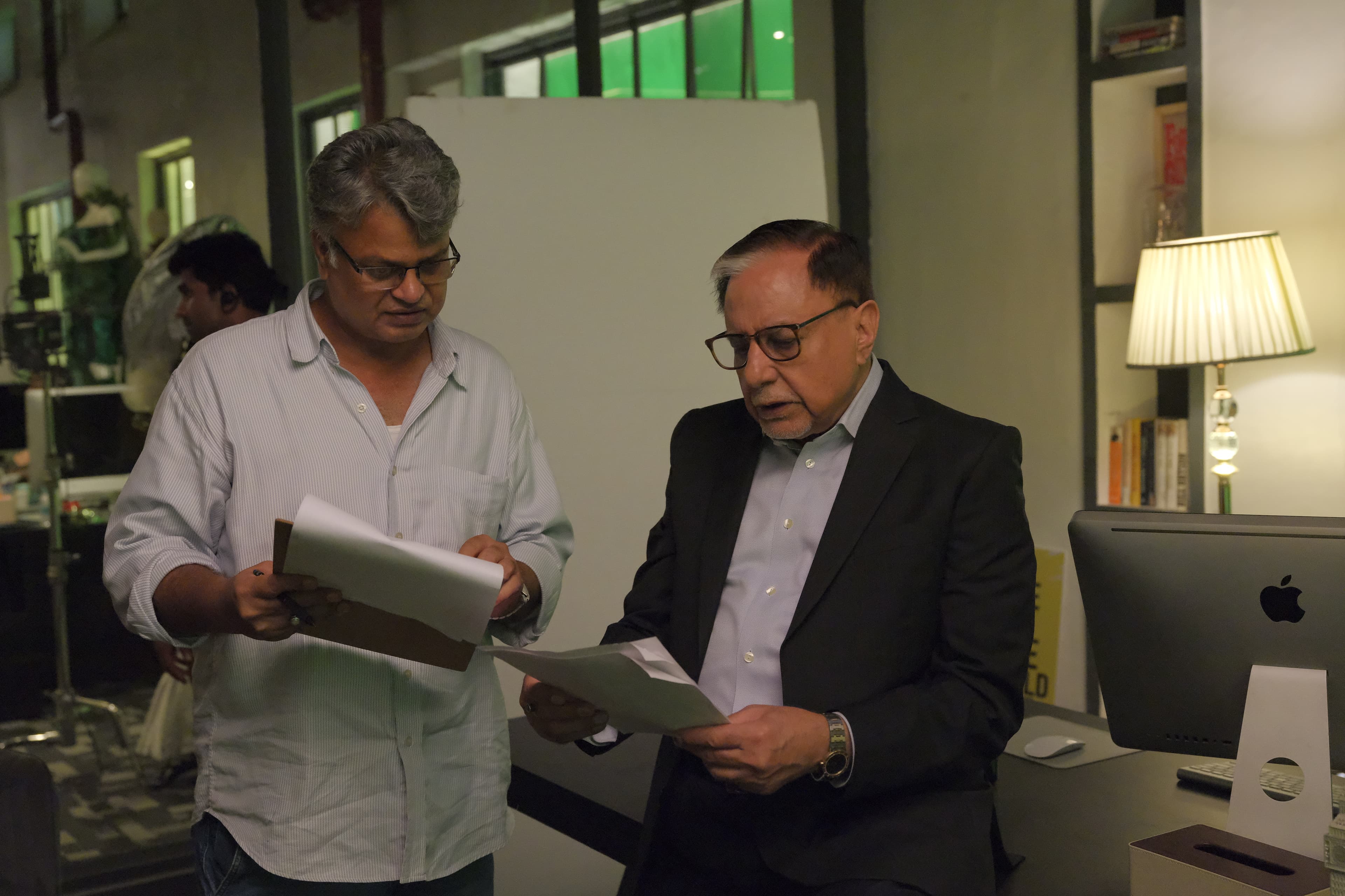Two men reading script pages together on a lit interior set